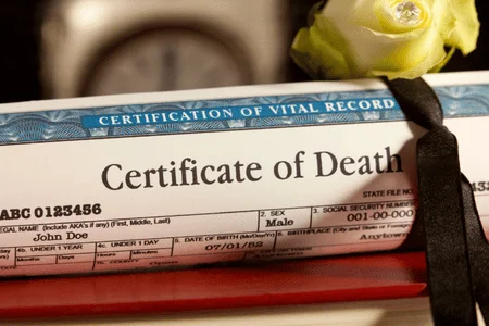 Death Certificate A rolled-up document on top of a wooden desk with the words “Certification of Vital Record” on the top and “Certificate of Death” beneath that. A yellow rose and clock sit behind the document.
